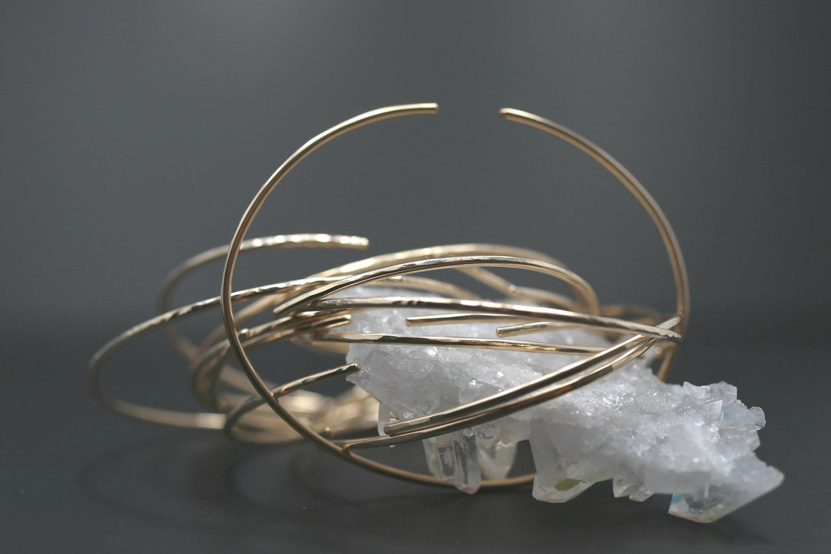 A large stack of thin yellow gold cuff bracelets is displayed over a white crystal cluster against a dark background.
