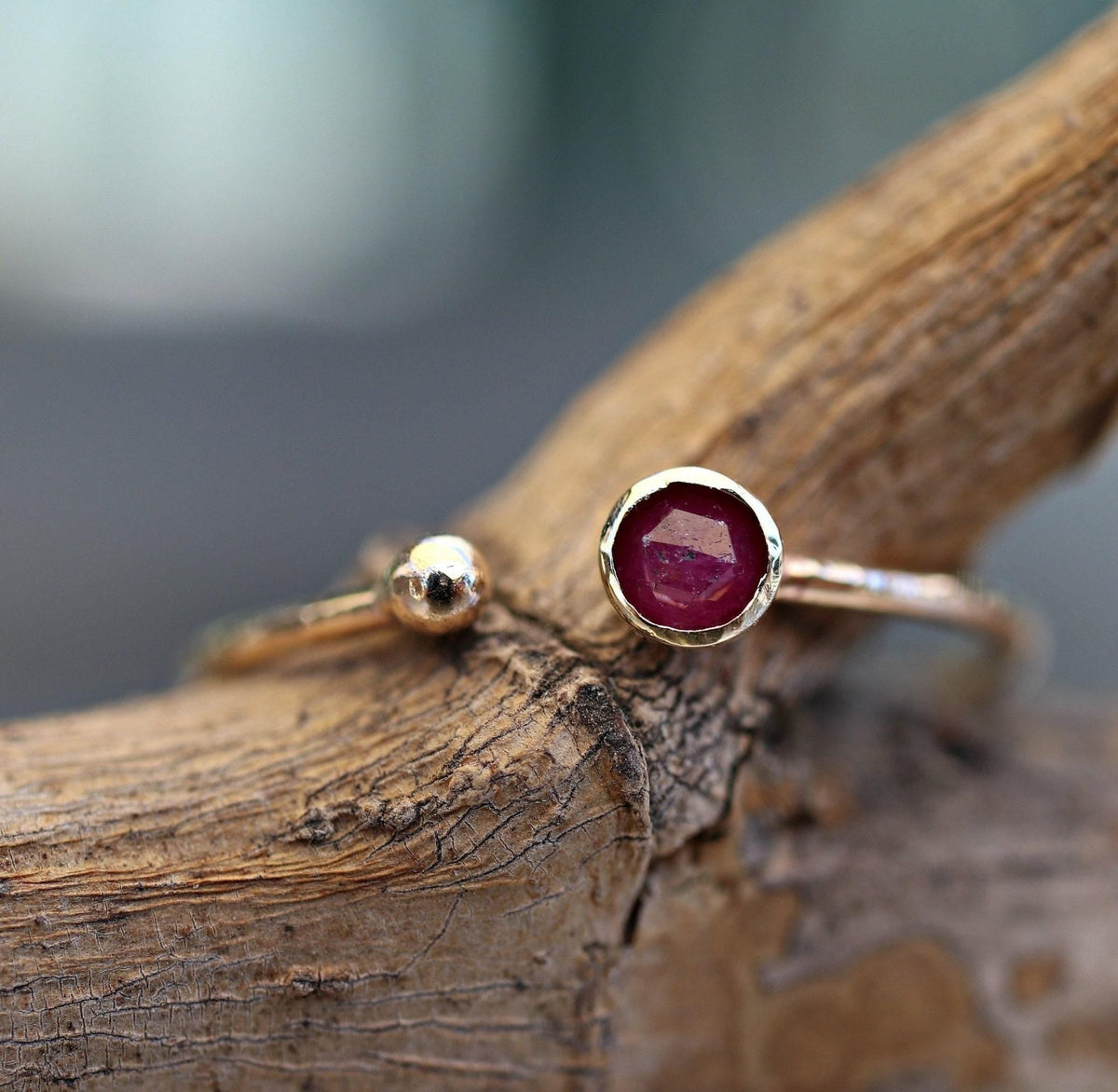 A gold open-cuff ring featuring a bezel-set faceted dark pink ruby on one end and a small gold ball on the other, resting on a textured wooden surface.