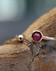 A gold open-cuff ring featuring a bezel-set faceted dark pink ruby on one end and a small gold ball on the other, resting on a textured wooden surface.