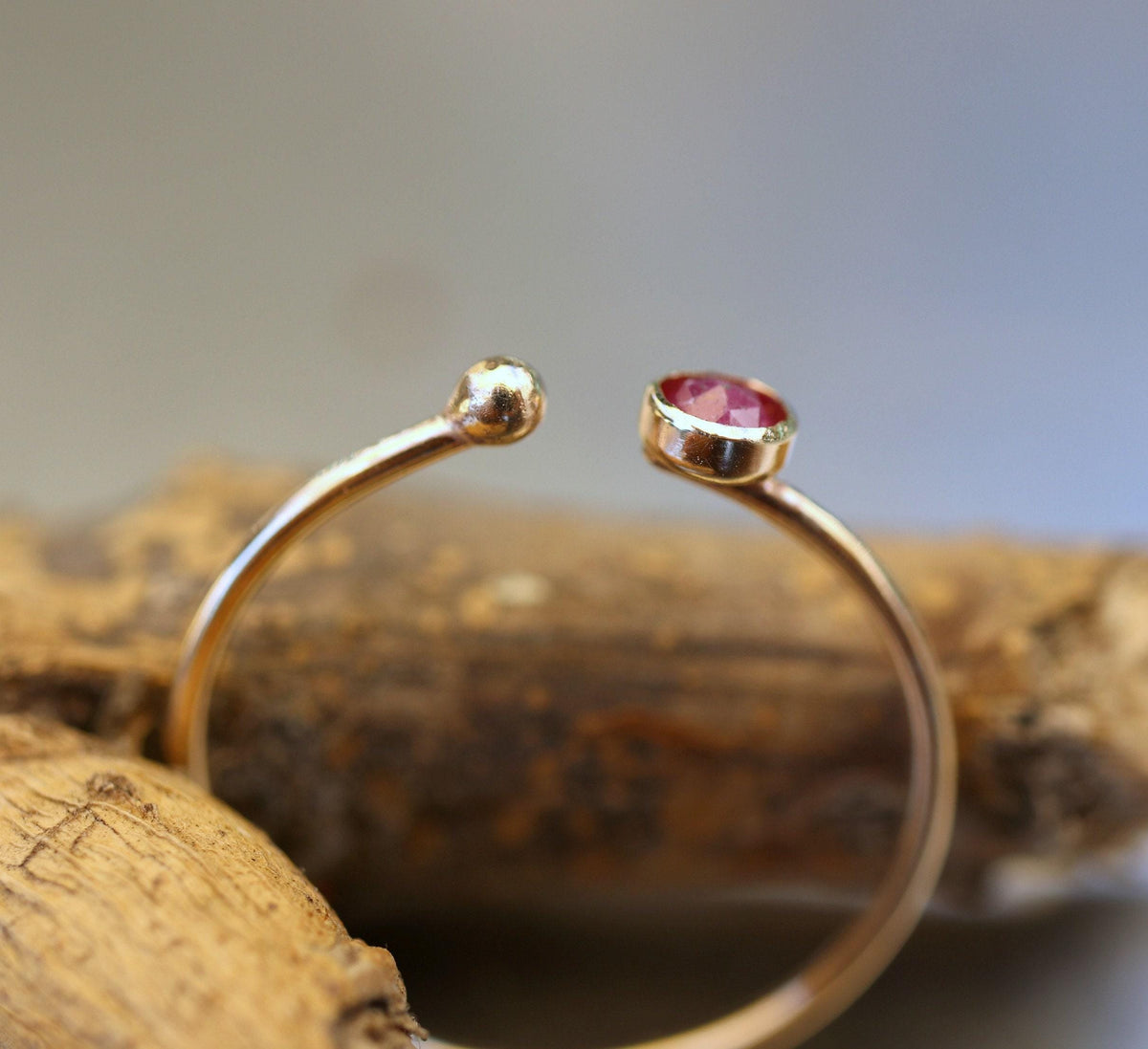 Close-up view of the gold open ring resting on wood, showing the gap between the bezel-set ruby and the terminal gold ball.