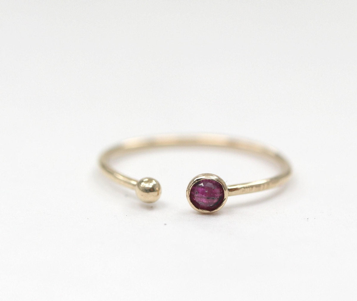 An open gold ring featuring a bezel-set ruby and a small ball end, placed on a plain white surface.