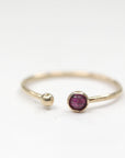 An open gold ring featuring a bezel-set ruby and a small ball end, placed on a plain white surface.