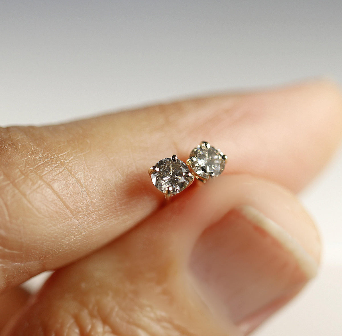 Two speckled grey diamond stud earrings held gently on the tip of a finger.