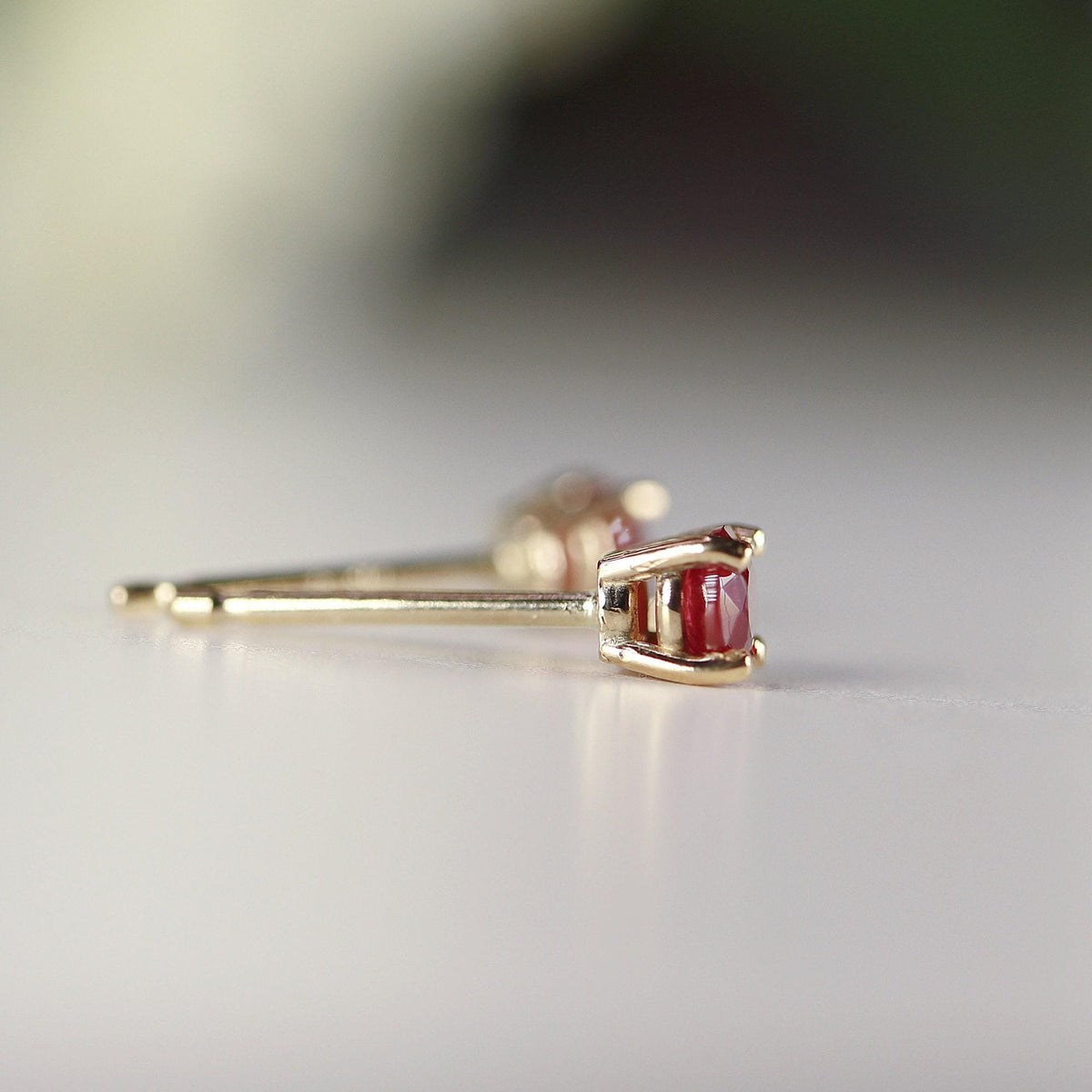 Side profile view of a ruby stud earring resting on a white surface, showing the gold post and prong setting.