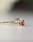 Side profile view of a ruby stud earring resting on a white surface, showing the gold post and prong setting.