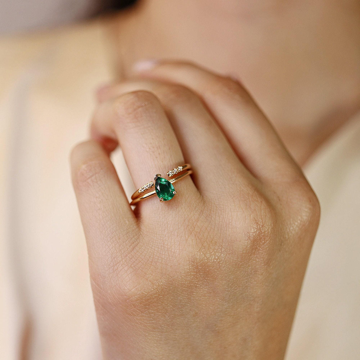 A pear-cut green emerald ring in a thin gold band is worn on a hand, paired with a delicate diamond band.