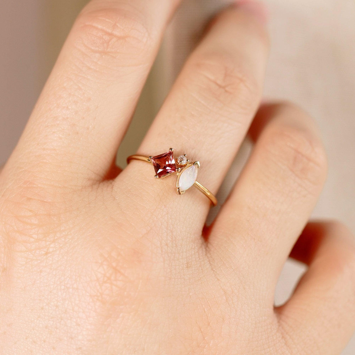 A gold ring with a square pink tourmaline, a marquise opal, and a small round diamond worn on a finger.