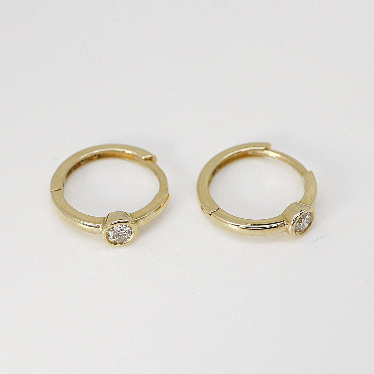 Two small gold hoop earrings with solitaire bezel-set diamonds viewed at a slight angle on a light background.
