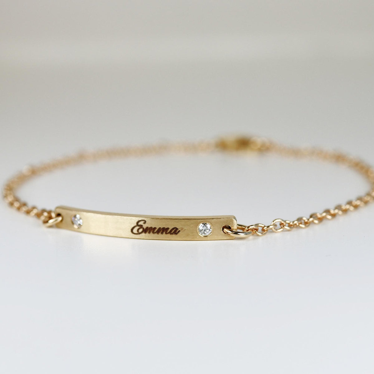 A close-up view of a gold chain bracelet with a curved bar engraved with Emma and flanked by two small sparkling stones.