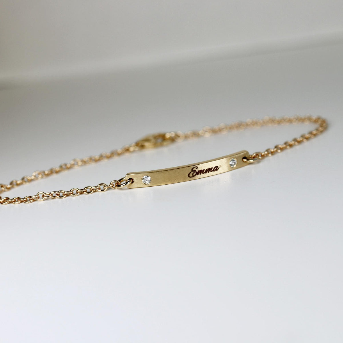 An angled shot of a thin gold chain bracelet showcasing a customized bar pendant with engraved text.