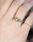 A hand wearing a thin gold ring with a green rectangular stone and a matching thin beaded diamond ring above it.