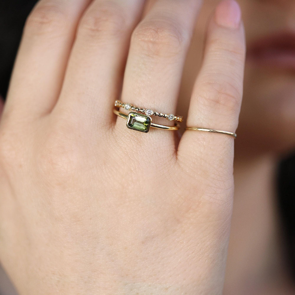 Close-up of a hand wearing a thin gold ring with a rectangular green gemstone and a delicate beaded diamond band stacked on top.