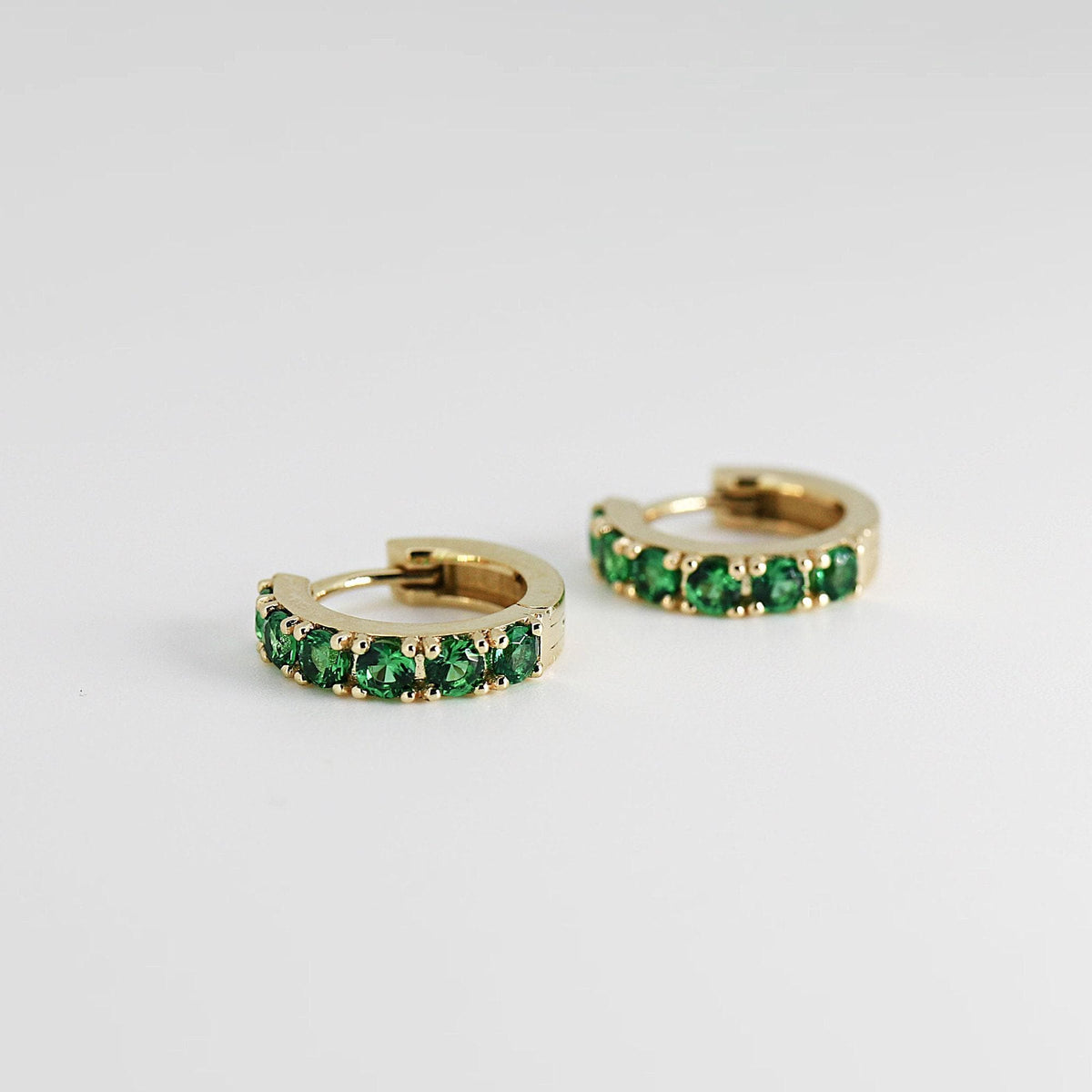 Front view of two gold hoop earrings set with round green gems, resting on a white surface.