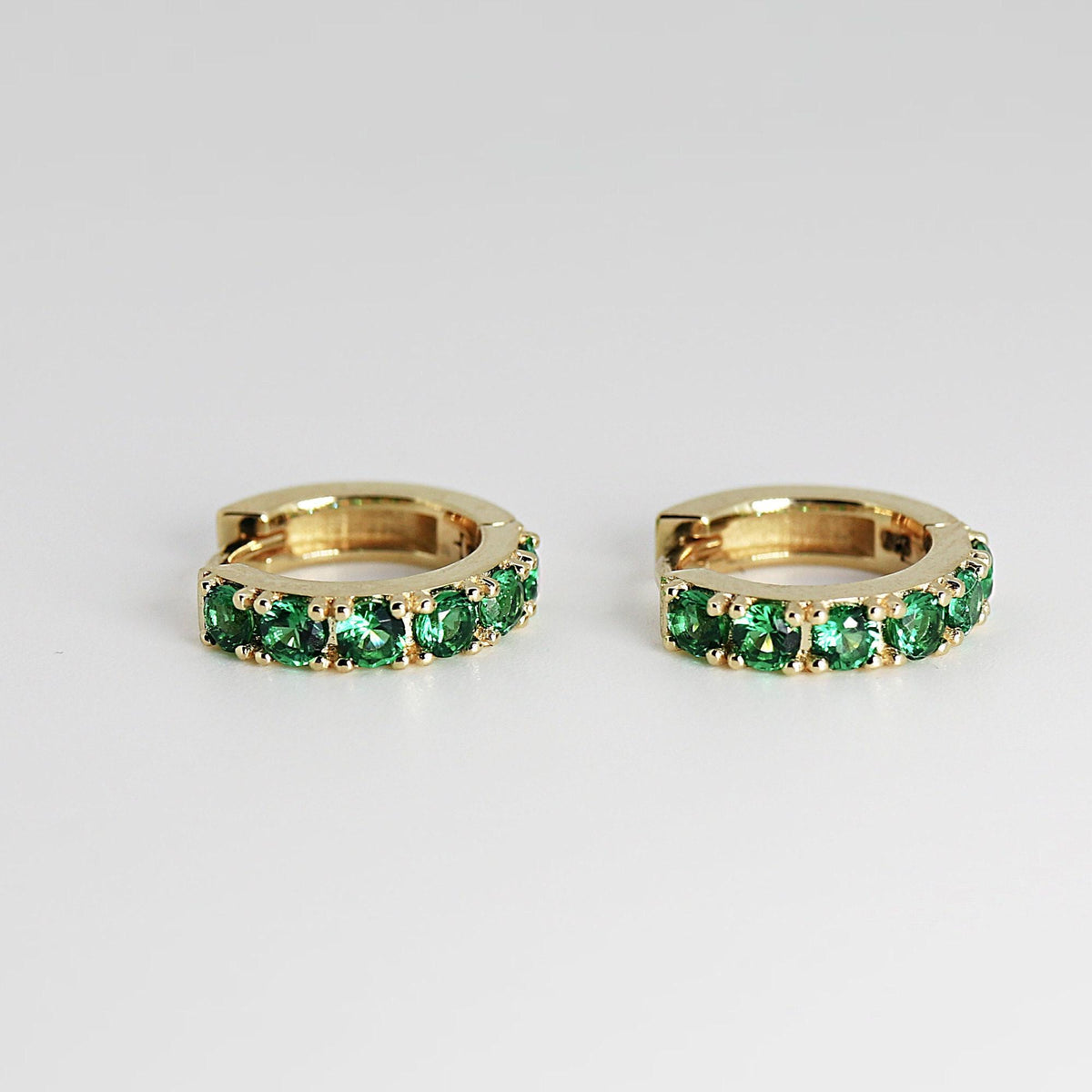 Close-up of two gold huggie hoop earrings lined with bright green stones, angled slightly toward each other.