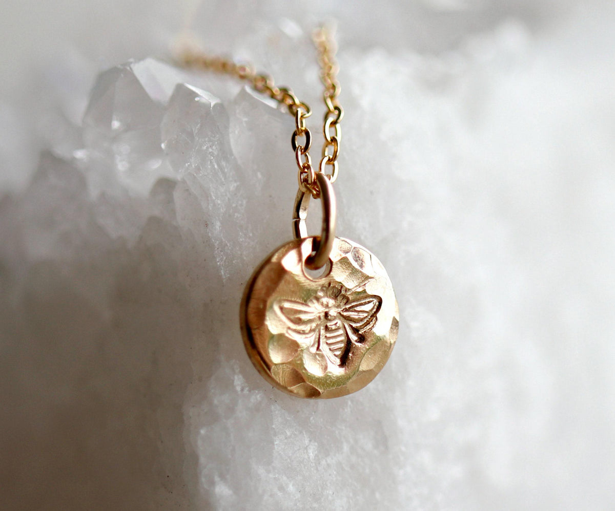 A close-up of a gold hammered coin pendant with an engraved bee motif, hanging from a delicate chain in front of a white crystal background.