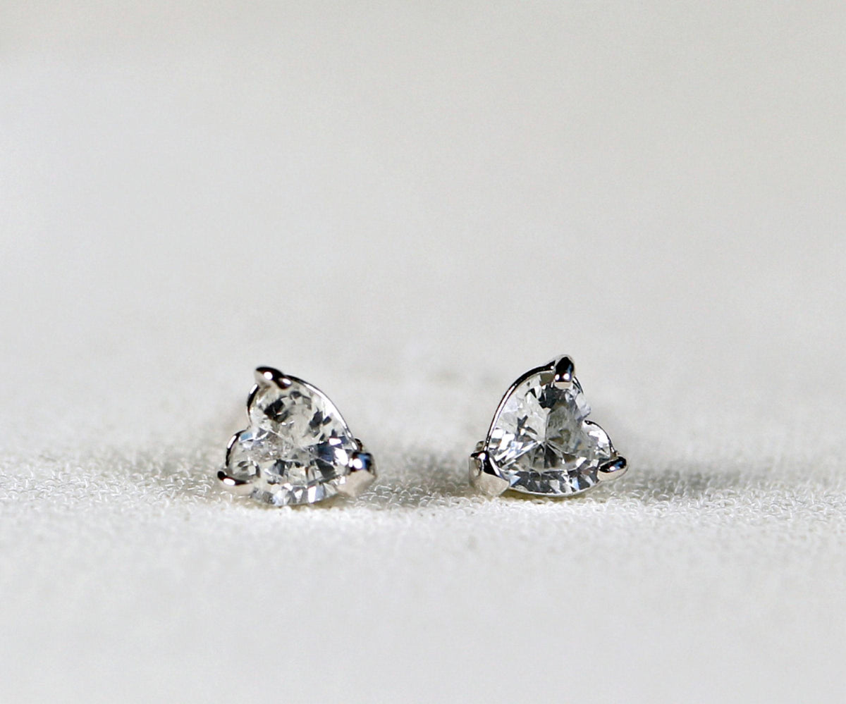 Two heart-cut white gemstone stud earrings resting on a textured white fabric surface.