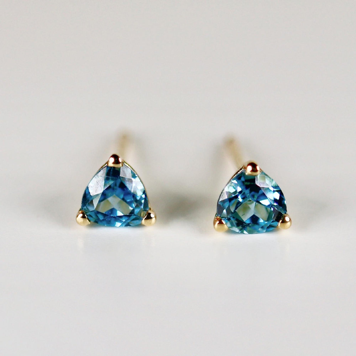 Front view of two trillion-cut blue gemstone stud earrings with gold prongs and posts on a white surface.