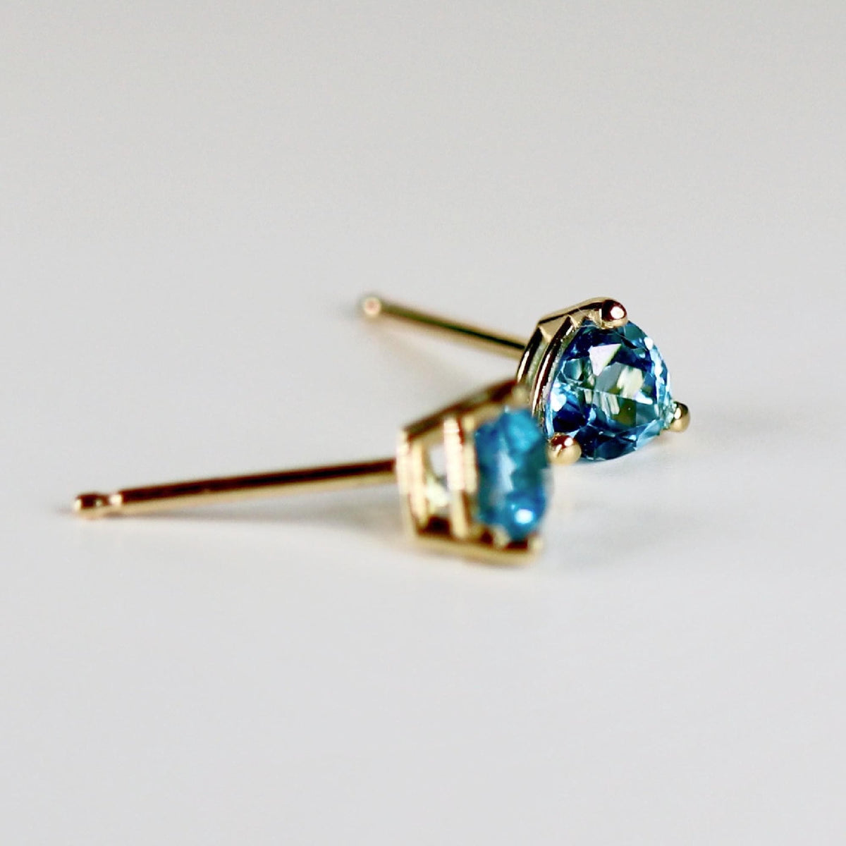 Angled view of two blue trillion-cut stud earrings showing the gold settings and posts.
