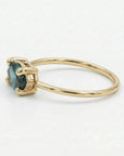 A close-up profile view of a gold ring featuring a prong-set blue-green gemstone on a simple band against a plain light background.