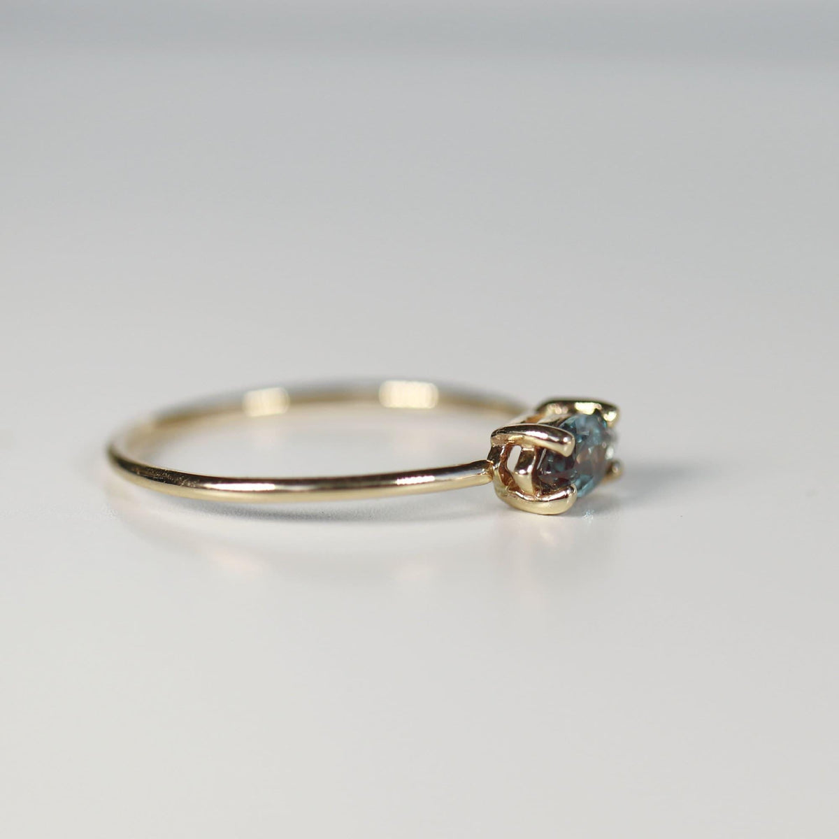 A side angle view of a thin gold ring showcasing the prong setting of a multi-toned gemstone resting on a white surface.