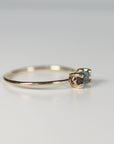 A side angle view of a thin gold ring showcasing the prong setting of a multi-toned gemstone resting on a white surface.
