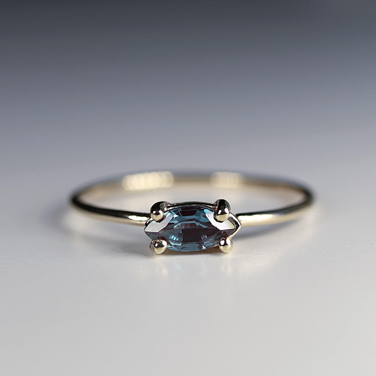 A front-angle view of a gold ring with a thin band and a horizontal marquise-cut blue gemstone in a four-prong setting on a pale background.