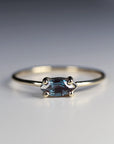 A front-angle view of a gold ring with a thin band and a horizontal marquise-cut blue gemstone in a four-prong setting on a pale background.