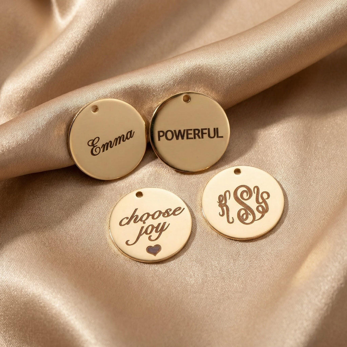 Four gold disc charms resting on satin fabric, engraved with Emma, POWERFUL, choose joy with a heart, and a monogram.
