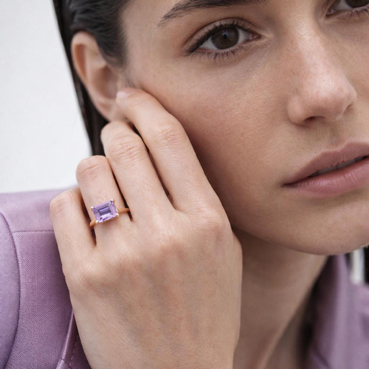 A close-up shot of a model wearing a gold ring with a rectangular purple gemstone on her hand, near her face.