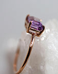 A side profile view of the gold ring with an emerald-cut purple gemstone, showing the prong setting and thin band against a crystal and light grey background.