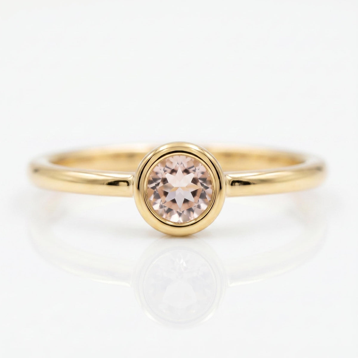 Front view of a gold ring featuring a bezel-set round light pink gemstone on a white background, casting a reflection.