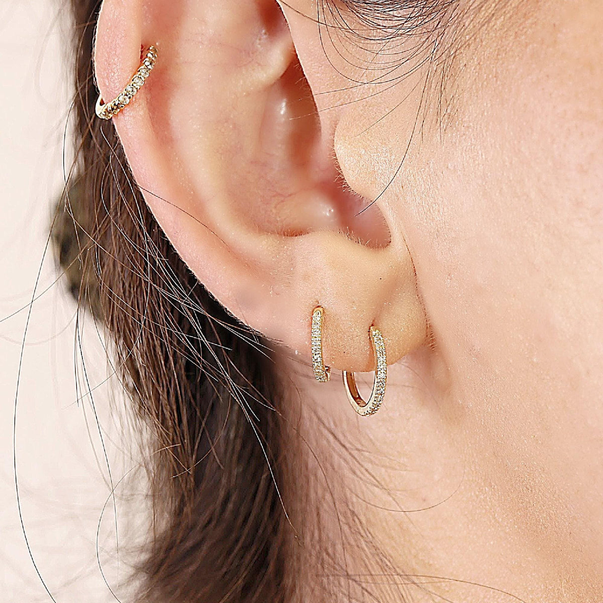 A close-up of an ear with a cartilage piercing and two small gold pave huggie earrings in the lobe.