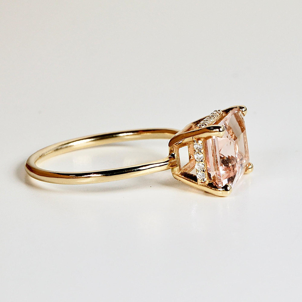 A gold ring with an emerald-cut pink stone rests on a white surface, viewed from the side to show the gallery and hidden halo diamonds.