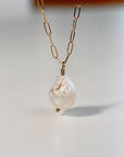 Detailed view of a large white baroque pearl hanging from a thin gold link chain against a neutral surface.