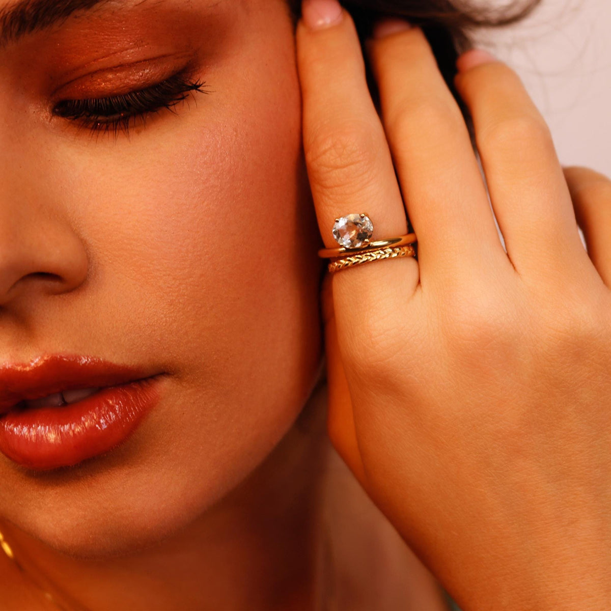A person wears a gold ring with a clear oval gemstone stacked with a braided gold band on their finger near their cheek.