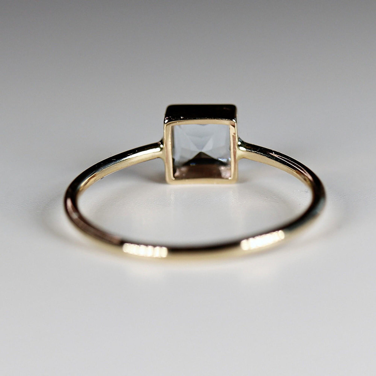 Rear view of a gold ring resting on a white surface with the open back of the square stone setting visible.