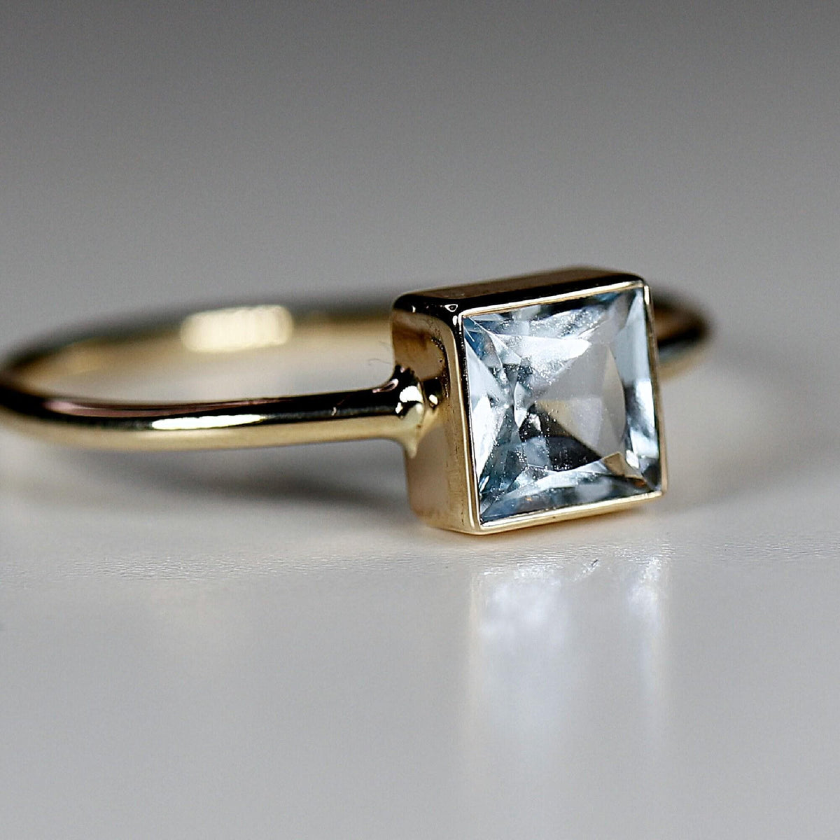 Macro side view of a gold bezel set square gemstone ring resting on a plain white surface.