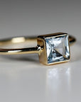 Macro side view of a gold bezel set square gemstone ring resting on a plain white surface.