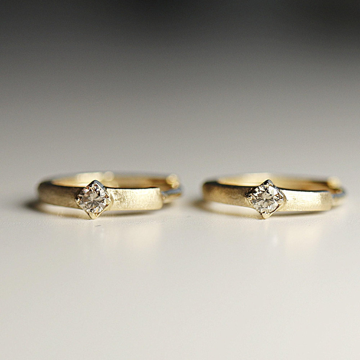 Two small gold hoop earrings with a brushed finish and a single square-set diamond on each lie upright on a white surface.