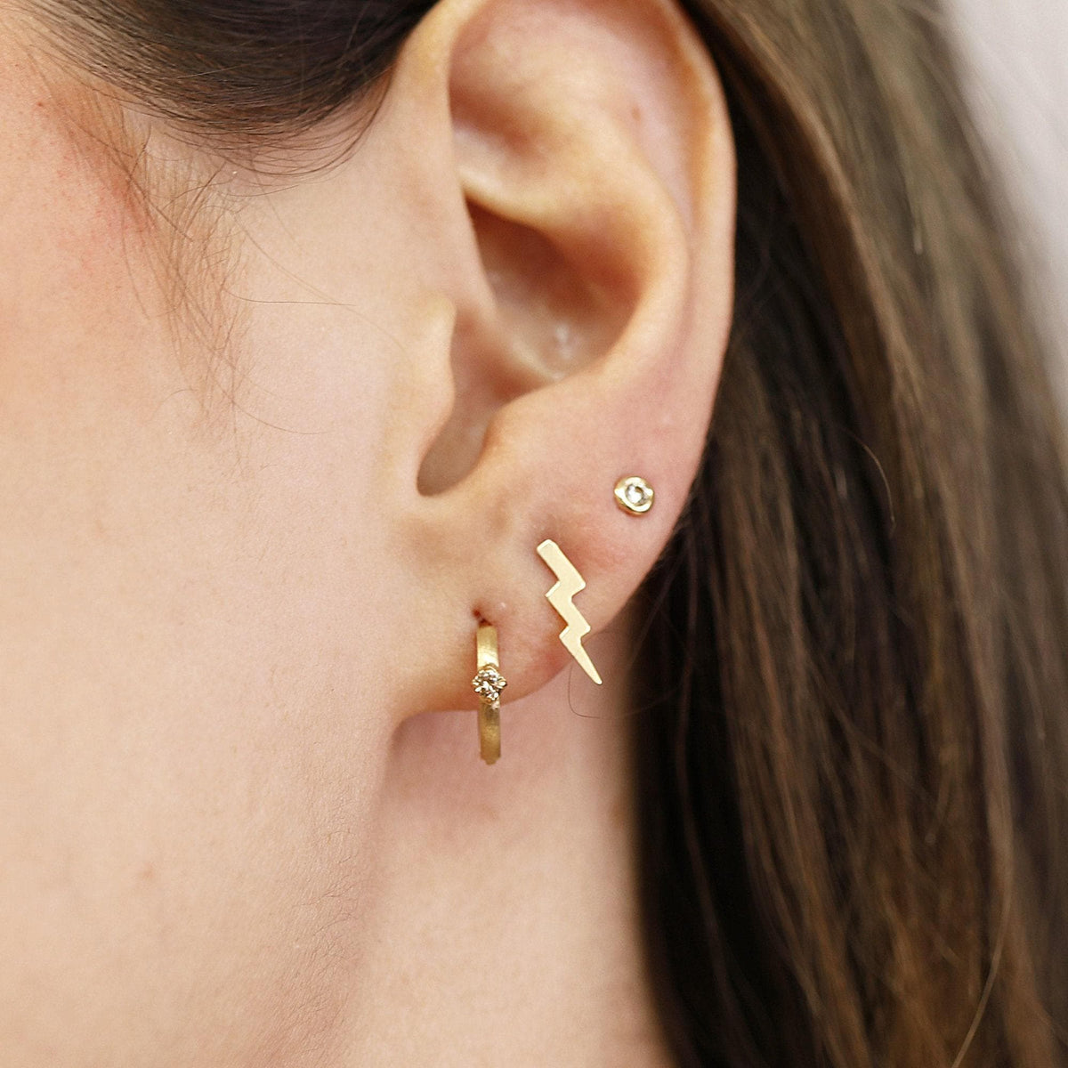 Close-up of an ear with three piercings wearing a small diamond huggie hoop, a gold lightning bolt stud, and a tiny stone stud.