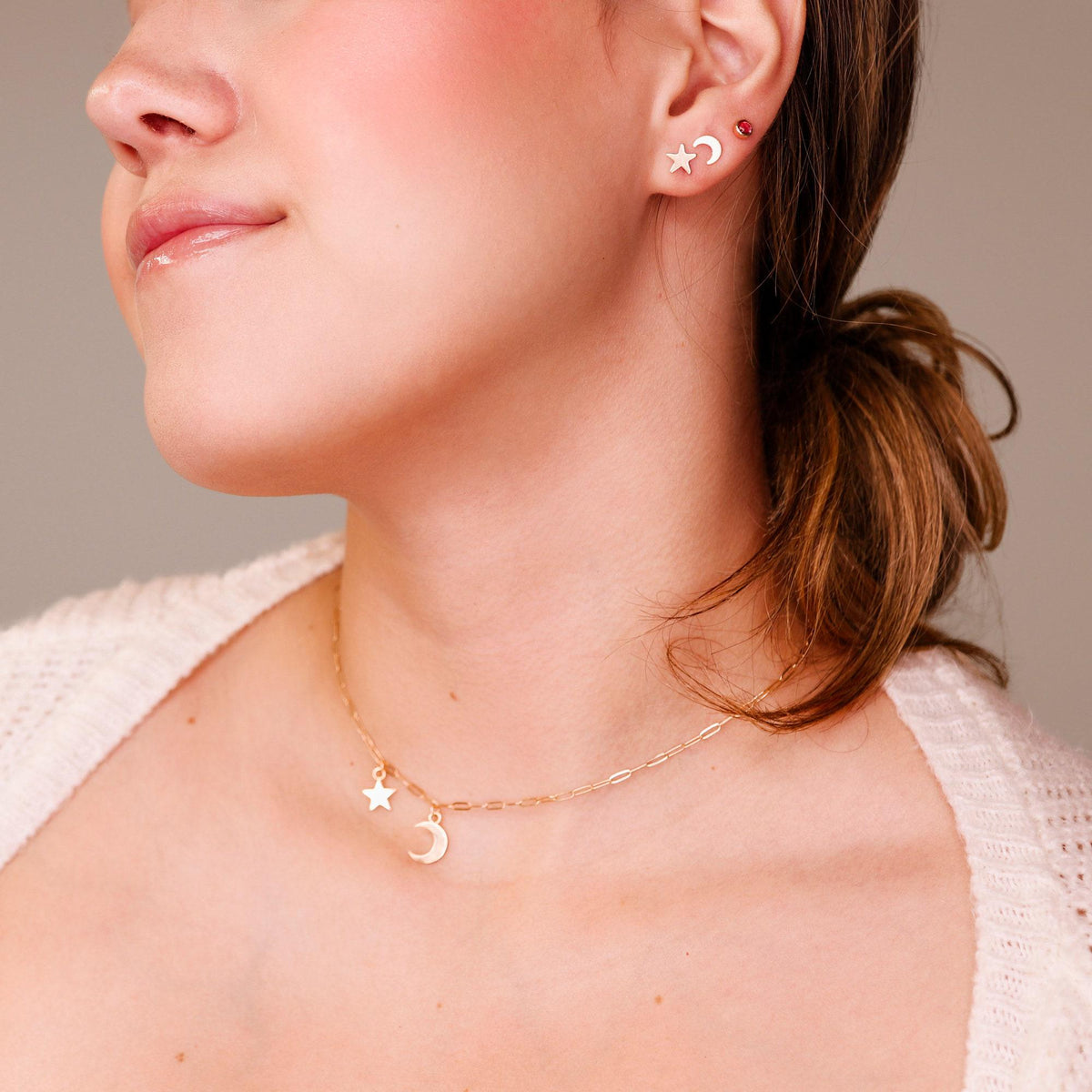 A woman wearing a gold star stud, a crescent moon stud, and a matching charm necklace.
