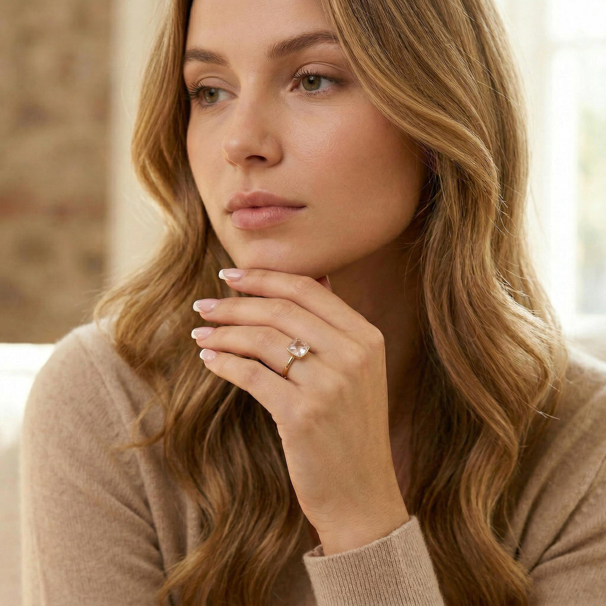 A woman with long wavy blonde hair resting her chin on her hand, wearing a cushion-cut pink gemstone ring.