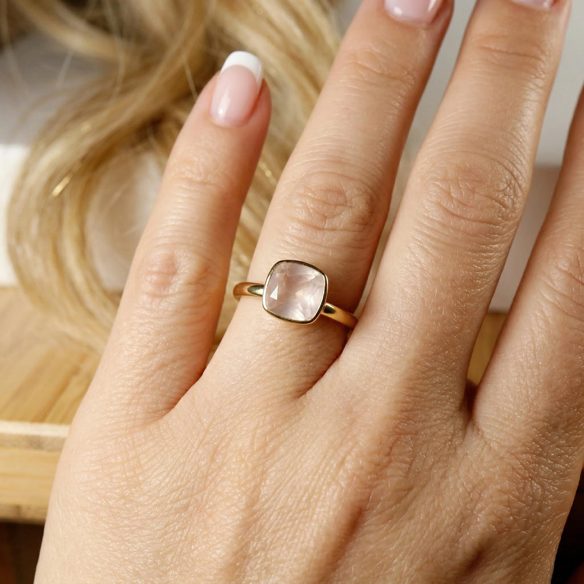 A close-up of a hand wearing a thin gold band with a bezel-set cushion-cut light pink stone.
