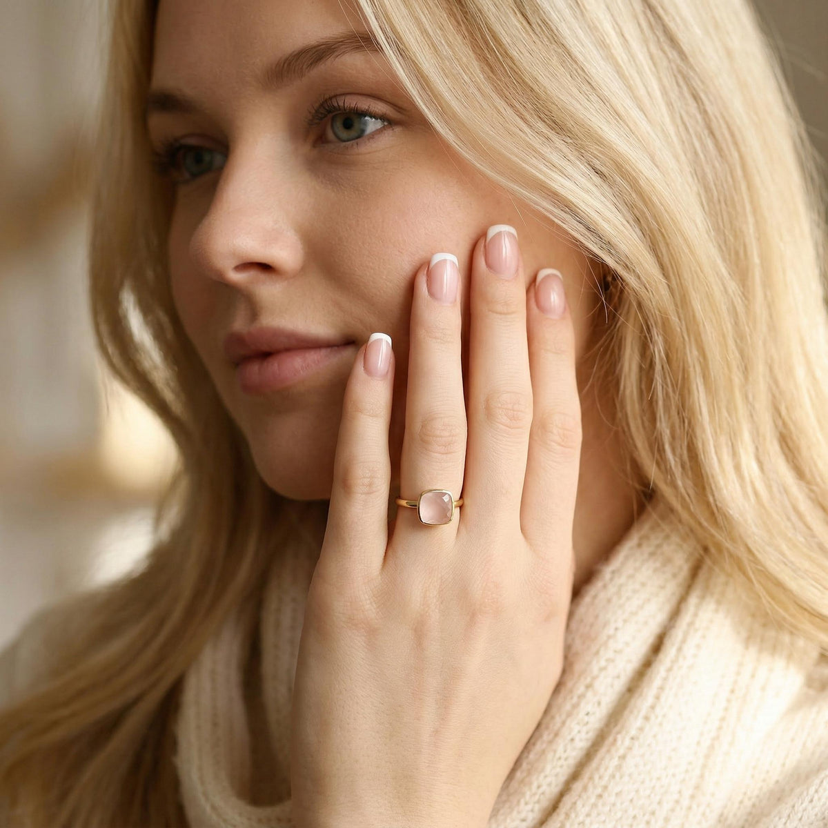 A blonde woman wearing a cream-colored knit sweater holds her hand up to her cheek, showcasing a gold ring with a pink stone.