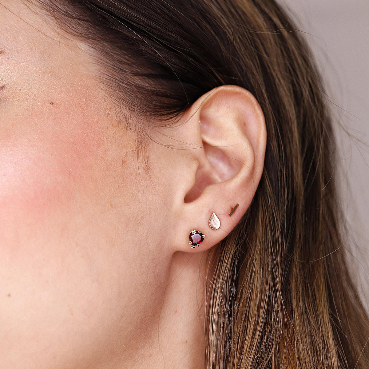 A side profile of an ear showcasing three different stud earrings including a red heart stone, a teardrop, and a tiny gold bar.