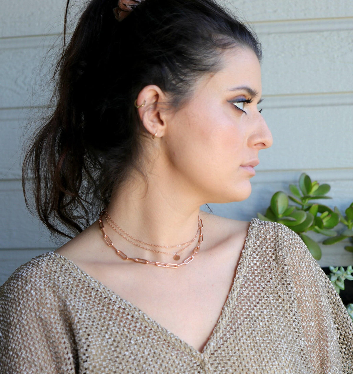 A person wearing a layered rose gold chain necklace featuring a chunky paperclip link strand and delicate chains with a small disc pendant.