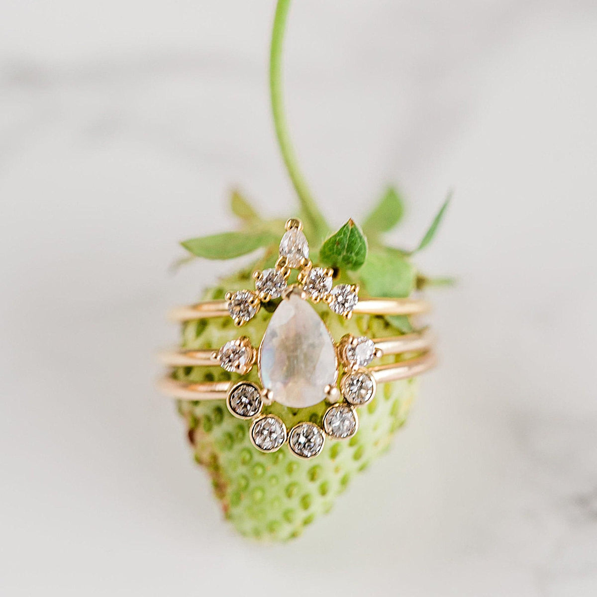A stacked set of gold gemstone rings rests on top of a fresh green strawberry.