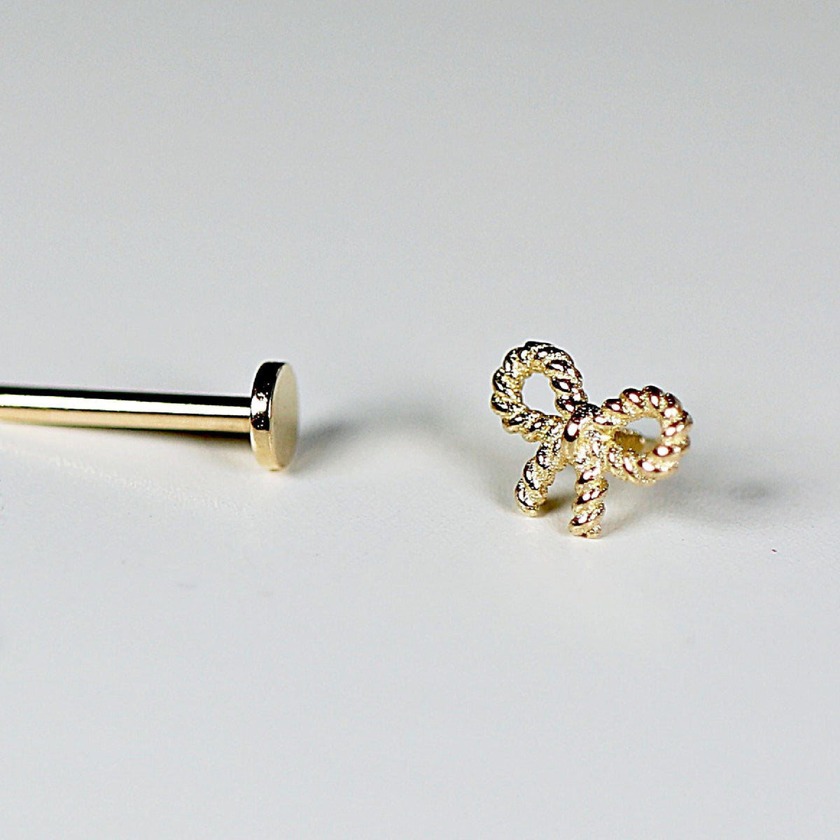 A gold bow piercing stud and its threaded flat back post are displayed separately on a light surface.