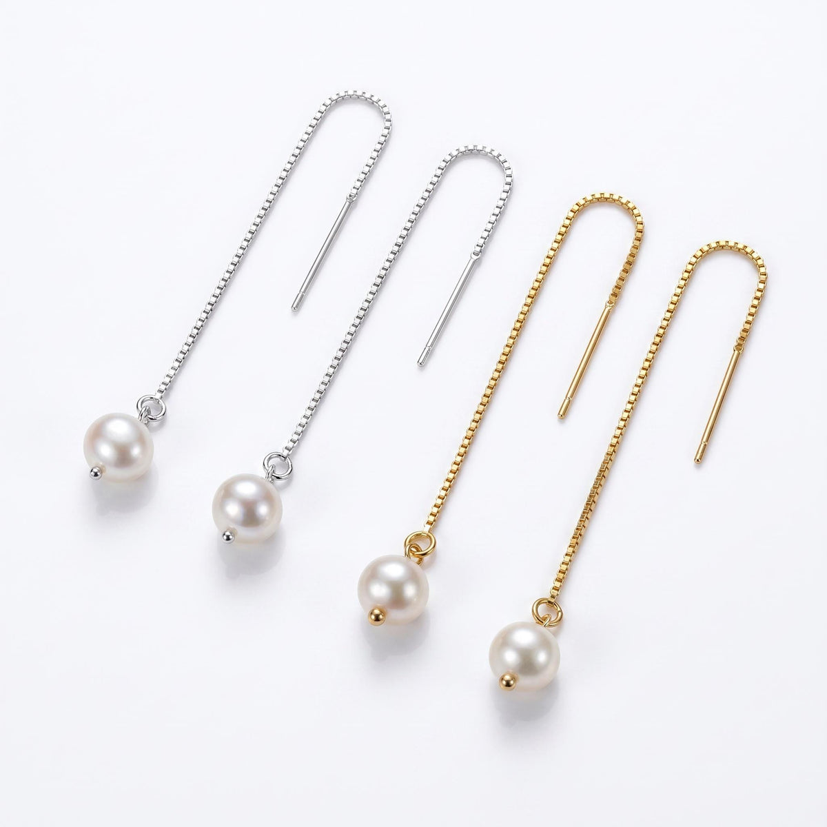Two pairs of threader earrings, one silver and one gold, each with a freshwater pearl drop, arranged on a white surface.