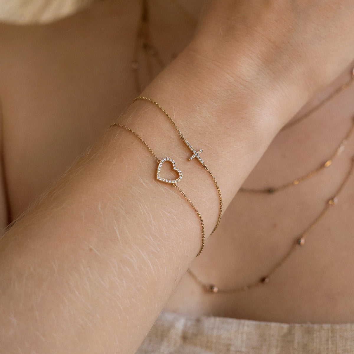 An angled view of a wrist showcasing a thin gold chain bracelet with a sparkling sideways cross paired with a heart bracelet.